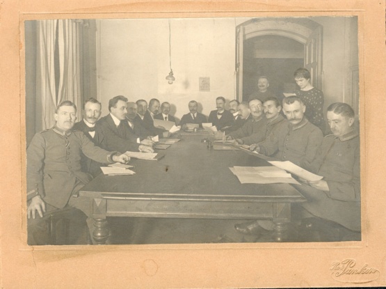 Arbeiter- und Soldatenrat Wismar © Quelle: Stadtgeschichtliches Museum Wismar II 17 Schwarzweiß Foto des Wismarer Arbeiter- und Soldatenrat: 15 Männer sitzen an einem rechteckigen Tisch, dahinter stehen ein weiterer Mann in einer geöffneten Tür und eine Frau