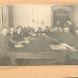 Schwarzweiß Foto des Wismarer Arbeiter- und Soldatenrat: 15 Männer sitzen an einem rechteckigen Tisch, dahinter stehen ein weiterer Mann in einer geöffneten Tür und eine Frau