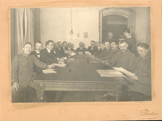 Schwarzweiß Foto des Wismarer Arbeiter- und Soldatenrat: 15 Männer sitzen an einem rechteckigen Tisch, dahinter stehen ein weiterer Mann in einer geöffneten Tür und eine Frau