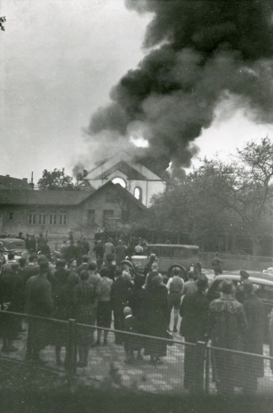Im Hintergrund Rauch und Flammen über der Synagoge, auf der Straße davor Menschen